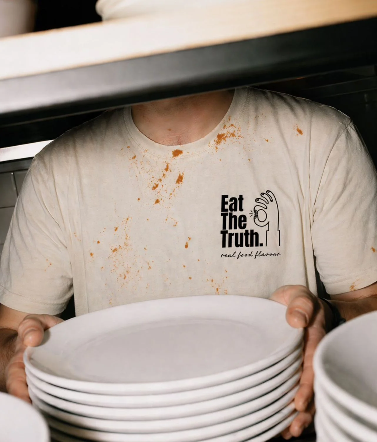 Chef in an Eat The Truth t-shirt holding a stack of plates in a professional kitchen