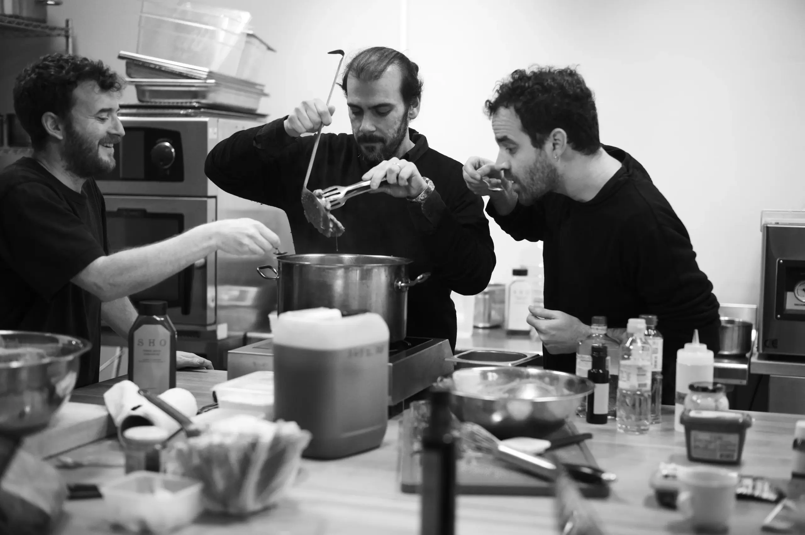 Trois chefs goûtant un bouillon directement à la louche au-dessus d'une grande marmite.