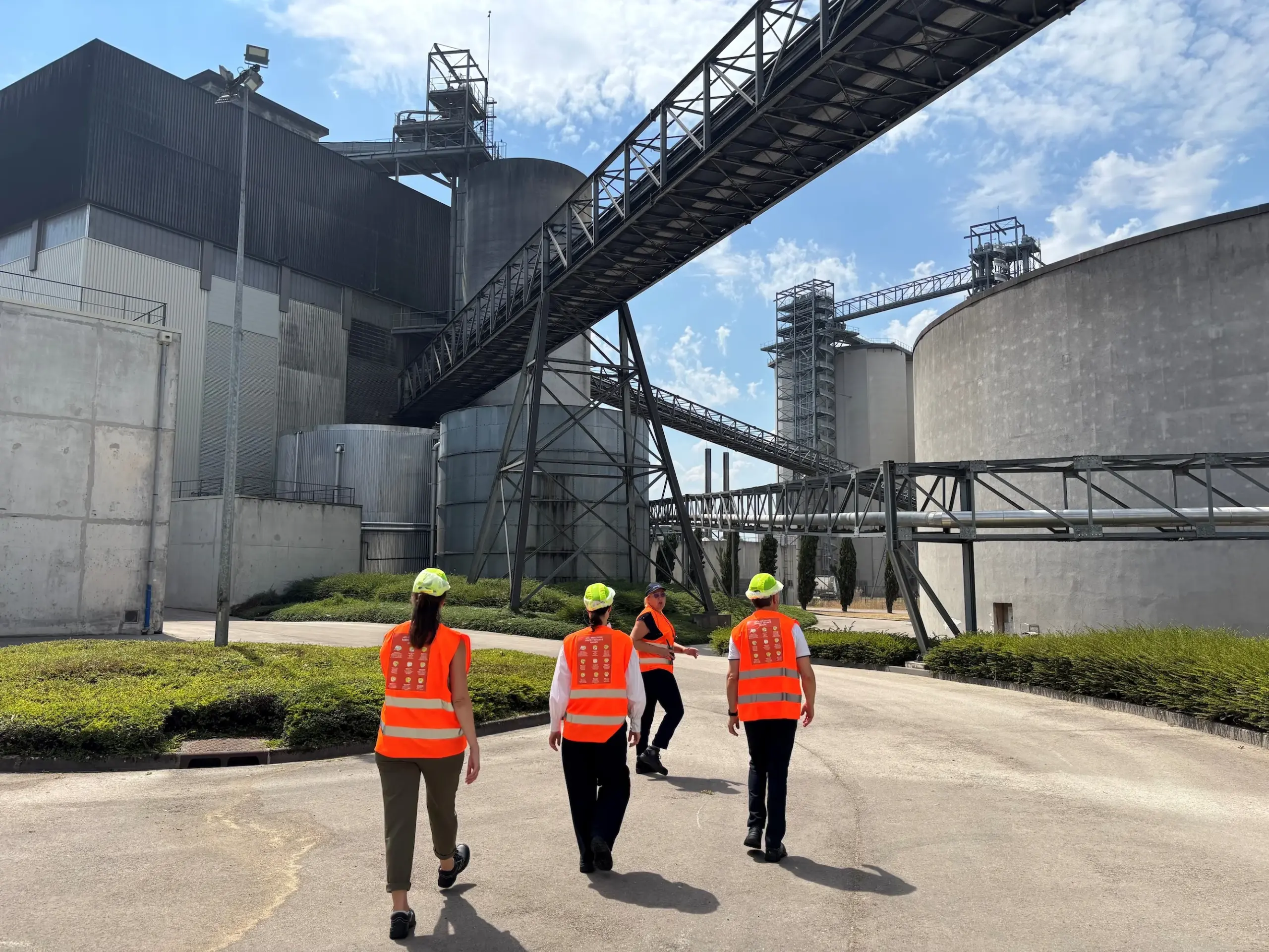 Team members wearing high-visibility orange vests and hard hats walking through a large-scale Soufflet Malt industrial production facility.
