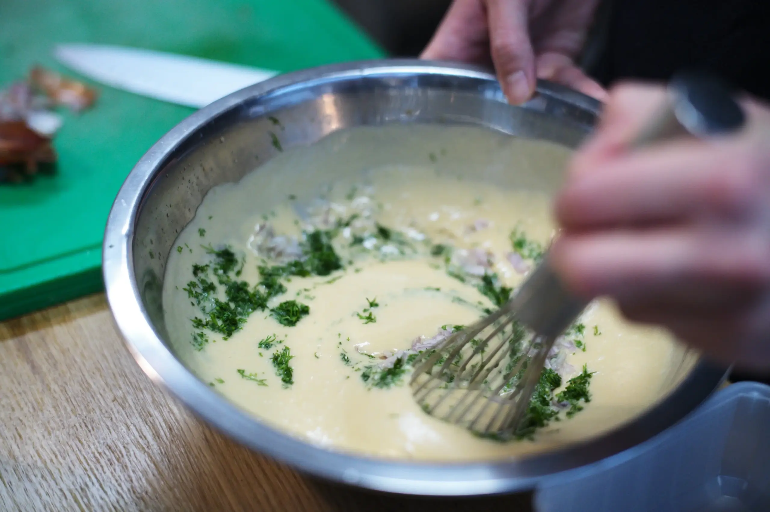 Whisking herbs into a rich creamy sauce base in a metal mixing bowl.